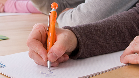Eine Hand schreibt etwas mit einem orangenen Kugelschreiber auf einen Block. (Link zu Informationen über den EHS-Fonds)
